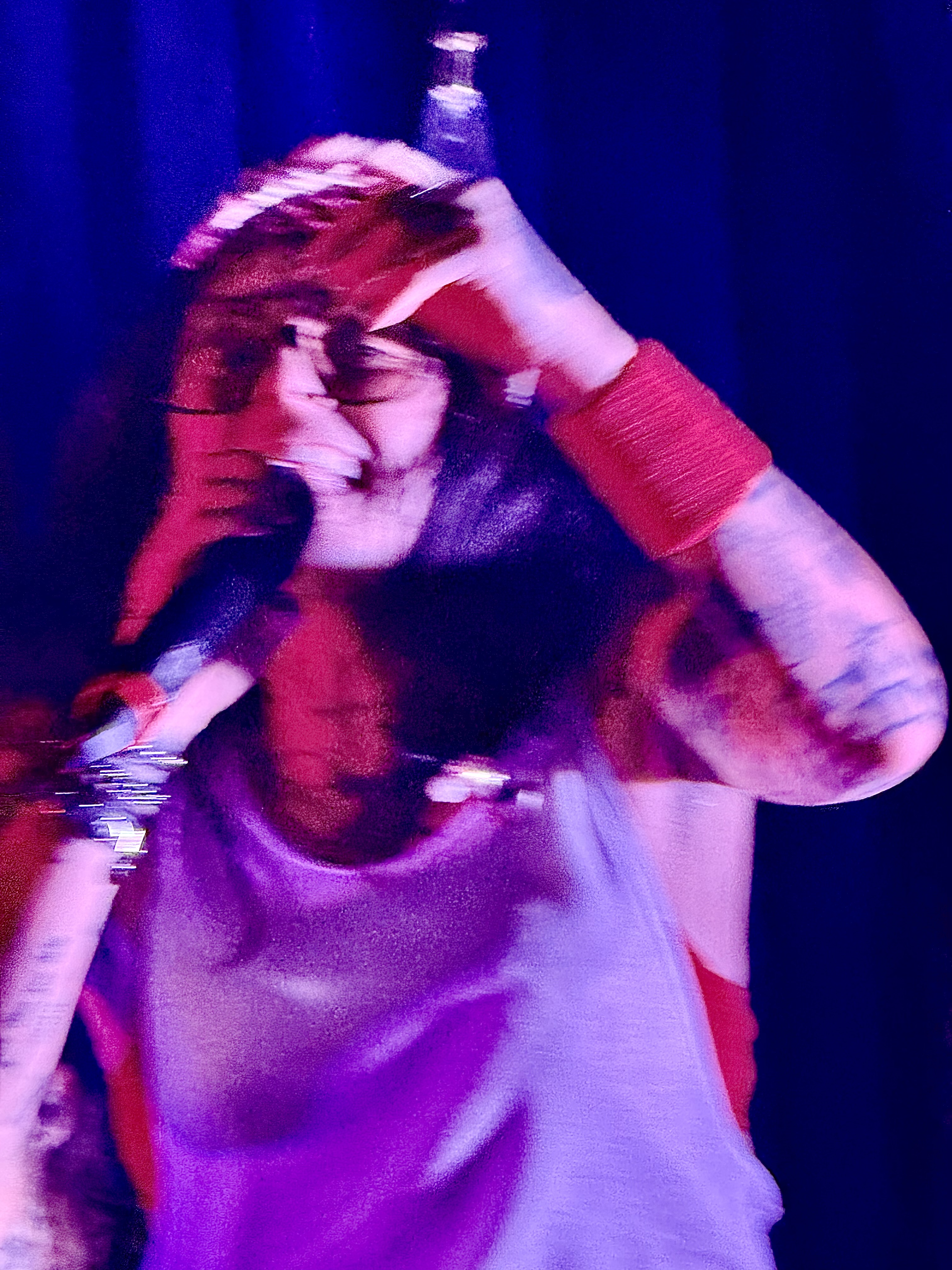 Singer holding a microphone with one hand raised to their head under purple and red stage lighting.