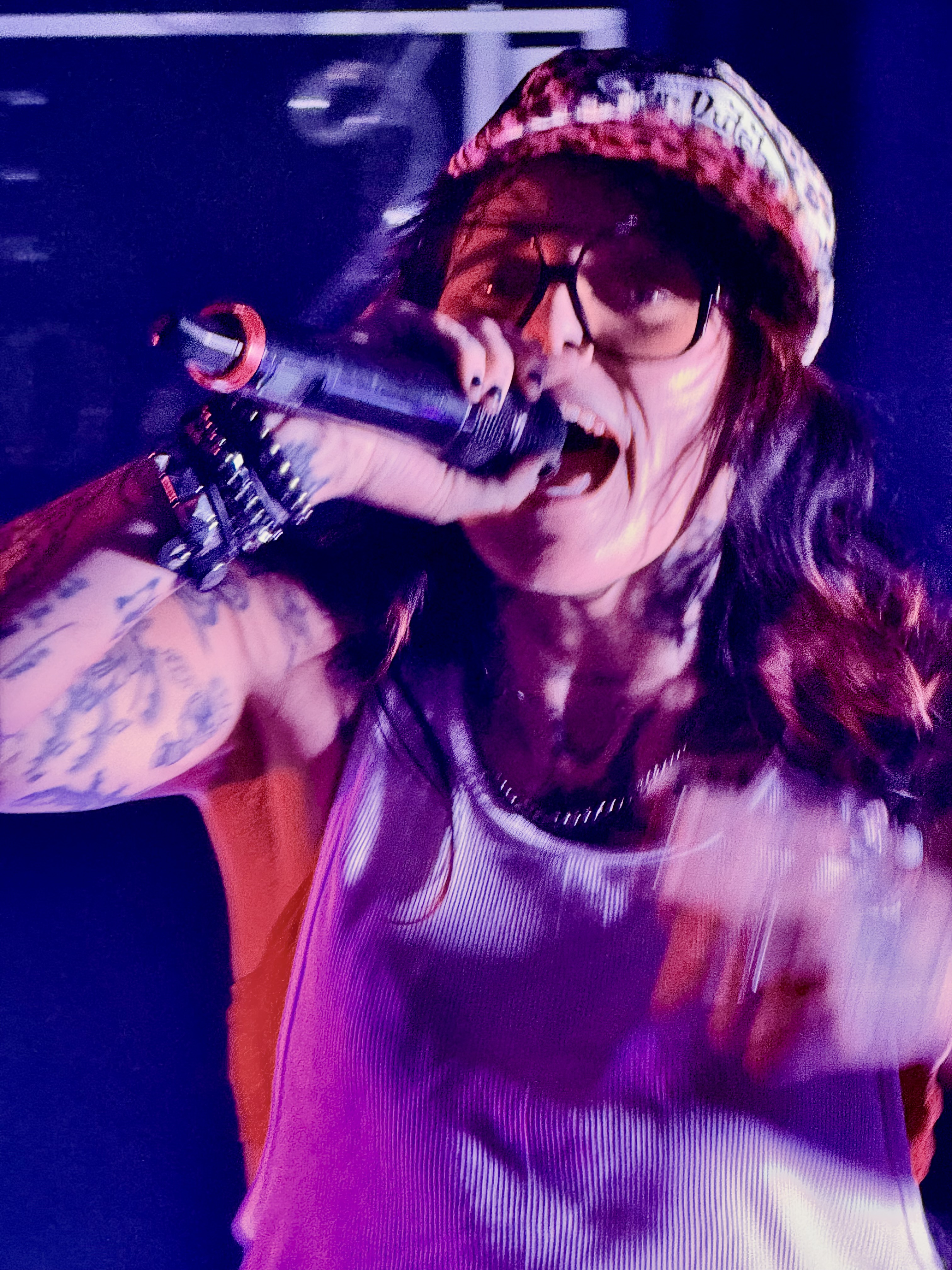 Close-up of a singer in sunglasses performing into a microphone under purple and red stage lighting.